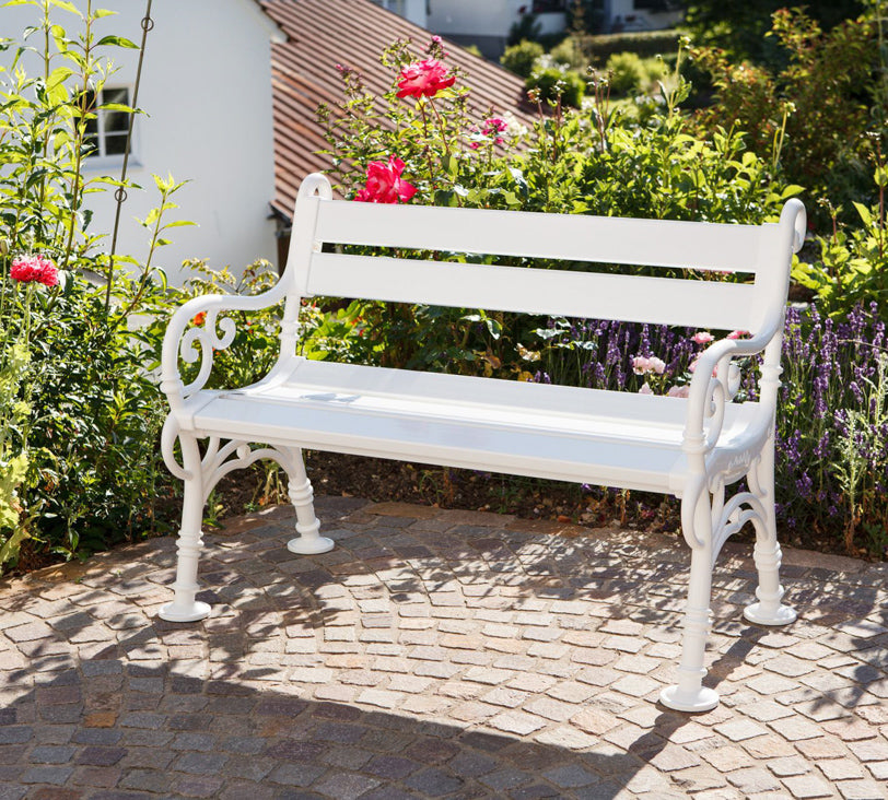Weiße Bank "Linderhof" als 2 Sitzer vor Blumenbeet in Gartenlandschaft, im Hintergrund unscharf erkennbare Häuser