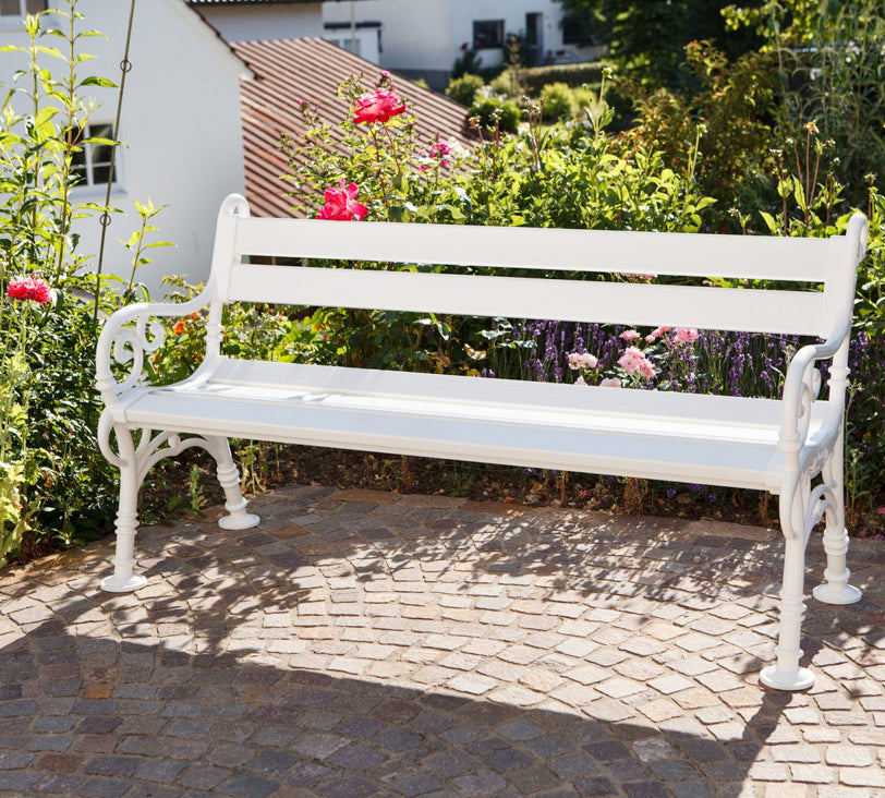 Weiße Bank "Linderhof" vor Blumenbeet in Gartenlandschaft, im Hintergrund unscharf erkennbare Häuser